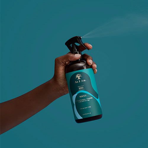 Hand holding a blue spray bottle labeled 'AS I AM' against a teal background